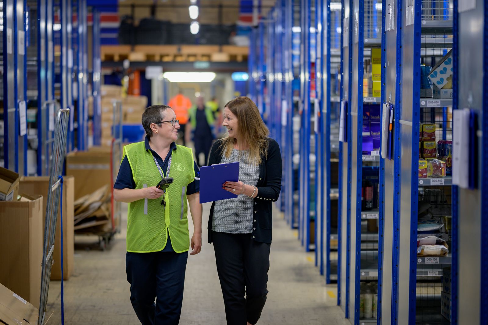 A supervisor with a clipboard walking a warehouse aisle alongside a worker in a hi-vis vest.